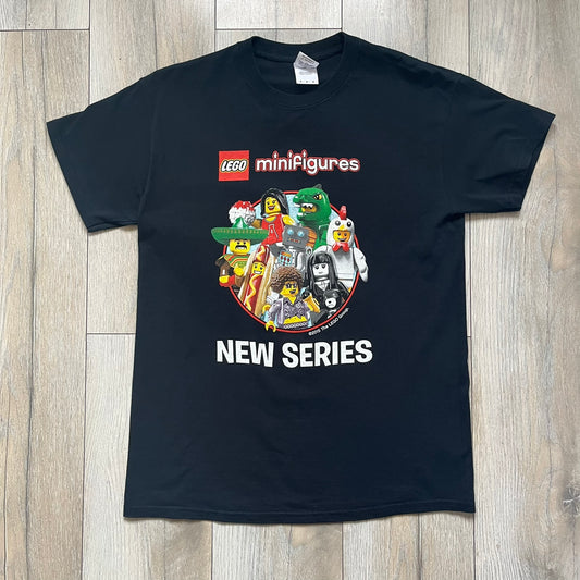 LEGO Minifigures “New Series” 2015 Tee is a black T-shirt featuring front and back prints. The front displays “LEGO Minifigures” text above eight characters arranged in a circle, with 2015 LEGO licensing and “New Series” text below. The back repeats the logo with a yellow minifigure in the center and “ASK ME” text underneath, likely produced as an employee promotional item.