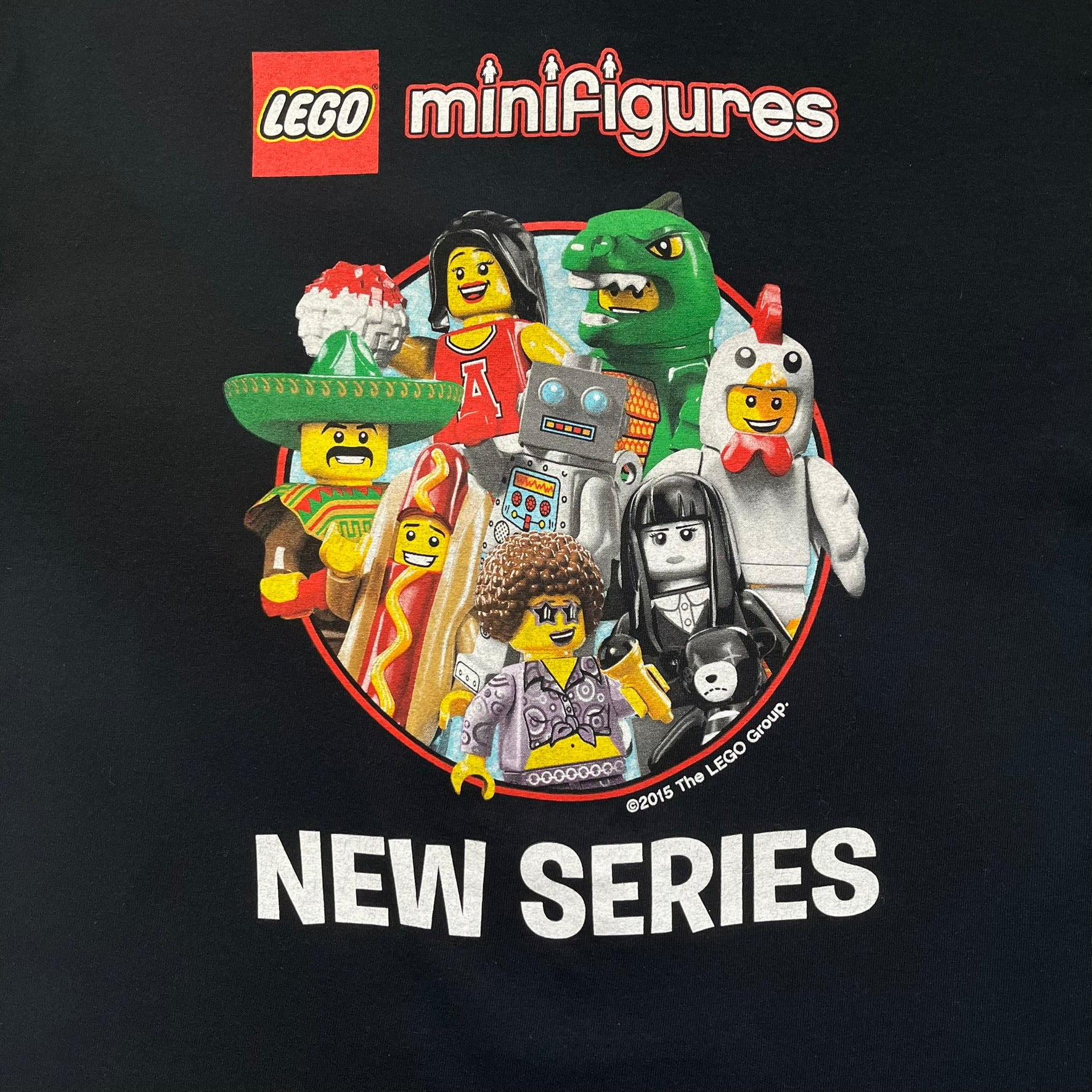 LEGO Minifigures “New Series” 2015 Tee is a black T-shirt featuring front and back prints. The front displays “LEGO Minifigures” text above eight characters arranged in a circle, with 2015 LEGO licensing and “New Series” text below. The back repeats the logo with a yellow minifigure in the center and “ASK ME” text underneath, likely produced as an employee promotional item.
