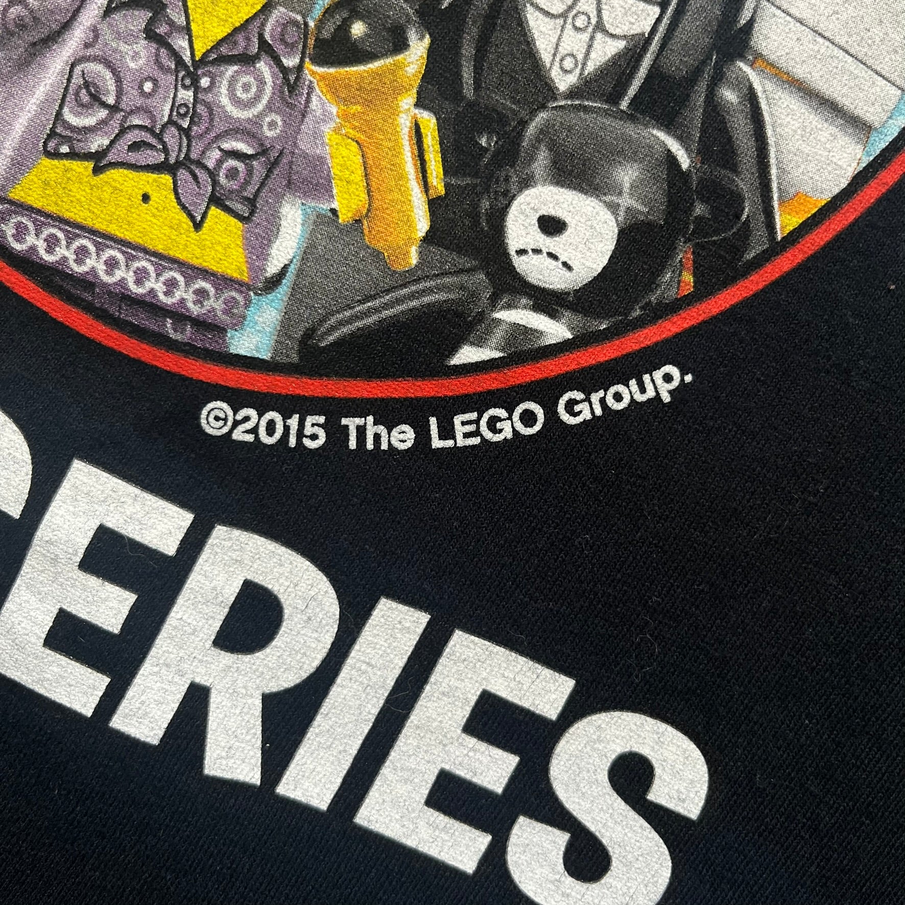 LEGO Minifigures “New Series” 2015 Tee is a black T-shirt featuring front and back prints. The front displays “LEGO Minifigures” text above eight characters arranged in a circle, with 2015 LEGO licensing and “New Series” text below. The back repeats the logo with a yellow minifigure in the center and “ASK ME” text underneath, likely produced as an employee promotional item.