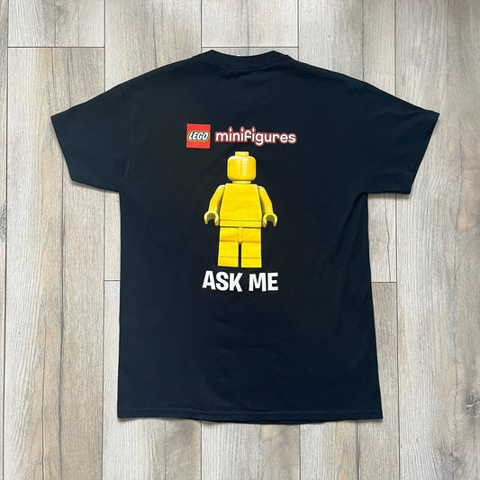 LEGO Minifigures “New Series” 2015 Tee is a black T-shirt featuring front and back prints. The front displays “LEGO Minifigures” text above eight characters arranged in a circle, with 2015 LEGO licensing and “New Series” text below. The back repeats the logo with a yellow minifigure in the center and “ASK ME” text underneath, likely produced as an employee promotional item.