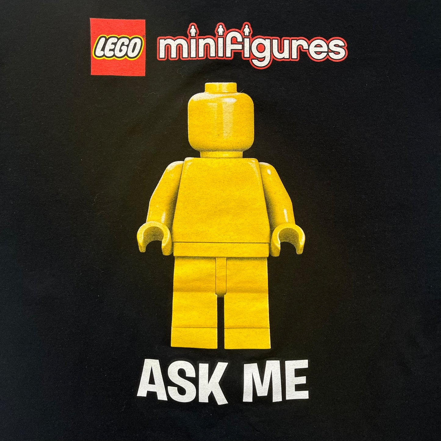 LEGO Minifigures “New Series” 2015 Tee is a black T-shirt featuring front and back prints. The front displays “LEGO Minifigures” text above eight characters arranged in a circle, with 2015 LEGO licensing and “New Series” text below. The back repeats the logo with a yellow minifigure in the center and “ASK ME” text underneath, likely produced as an employee promotional item.