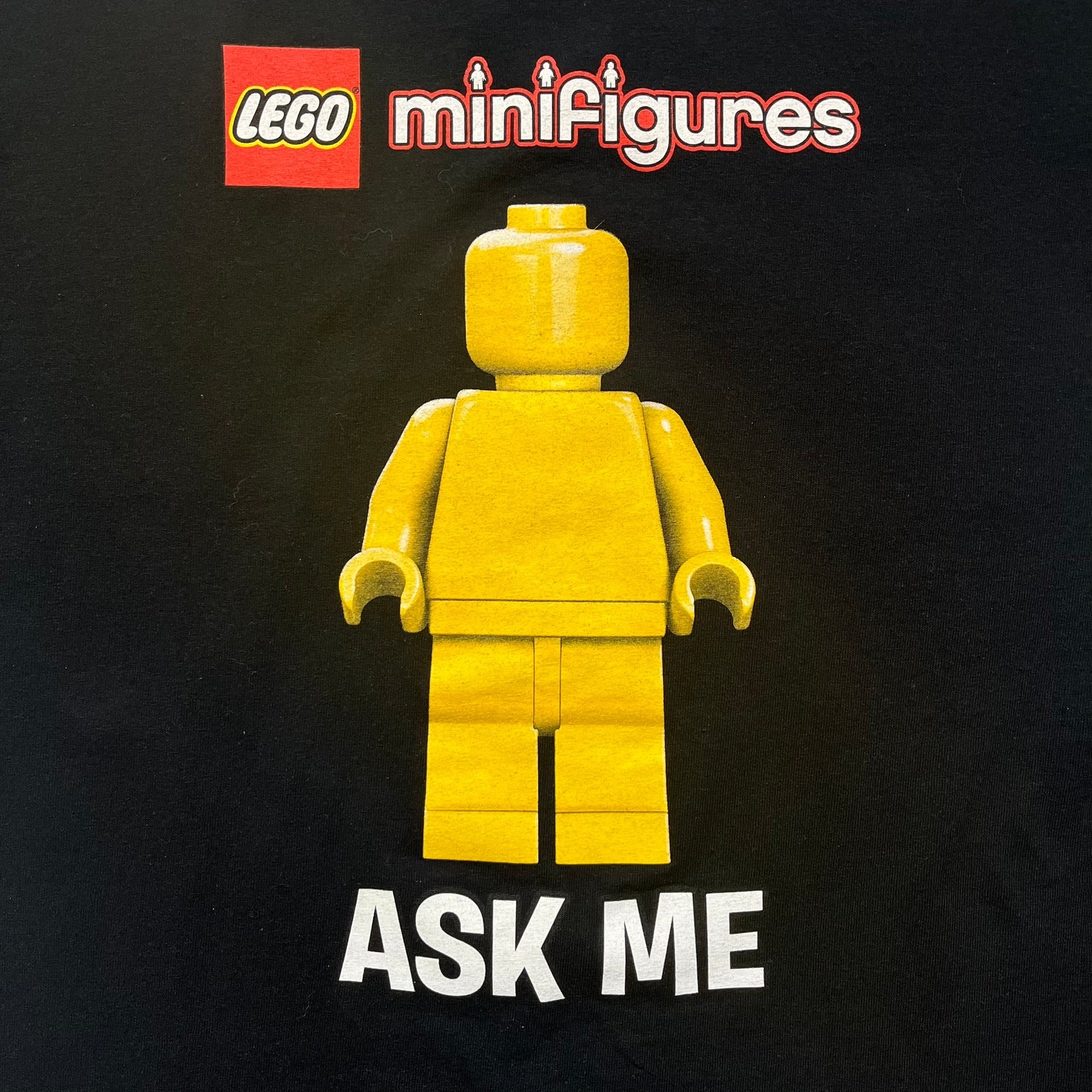 LEGO Minifigures “New Series” 2015 Tee is a black T-shirt featuring front and back prints. The front displays “LEGO Minifigures” text above eight characters arranged in a circle, with 2015 LEGO licensing and “New Series” text below. The back repeats the logo with a yellow minifigure in the center and “ASK ME” text underneath, likely produced as an employee promotional item.