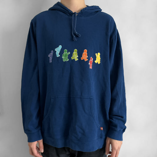 Lego Rainbow Minifigures Blue Hoodie from 2010, officially licensed by the Lego Group. Crafted from 100% cotton, it features seven colorful Lego minifigures in motion across the chest and a single red figure waving on the upper back. Designed in a bright blue colorway with a small Lego tag by the front pocket.