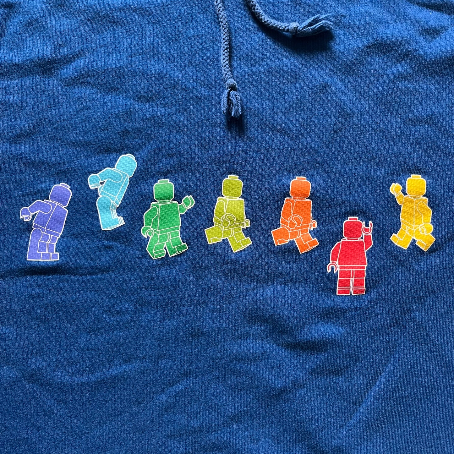 Lego Rainbow Minifigures Blue Hoodie from 2010, officially licensed by the Lego Group. Crafted from 100% cotton, it features seven colorful Lego minifigures in motion across the chest and a single red figure waving on the upper back. Designed in a bright blue colorway with a small Lego tag by the front pocket.