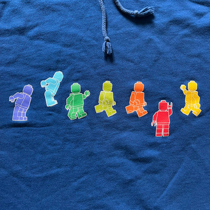 Lego Rainbow Minifigures Blue Hoodie from 2010, officially licensed by the Lego Group. Crafted from 100% cotton, it features seven colorful Lego minifigures in motion across the chest and a single red figure waving on the upper back. Designed in a bright blue colorway with a small Lego tag by the front pocket.