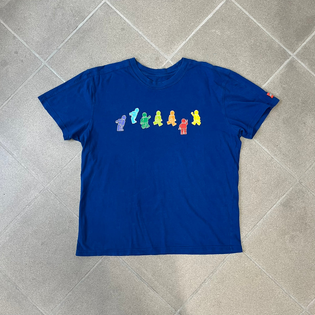 Lego Rainbow Minifigures Blue Tee from 2010, officially licensed by the Lego Group. Crafted from 100% cotton, it features seven colorful Lego minifigures in motion across the chest. Designed in a blue colorway with a small Lego tag on the left sleeve.