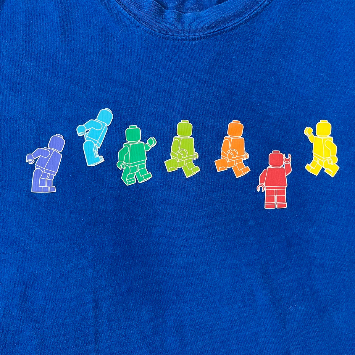 Lego Rainbow Minifigures Blue Tee from 2010, officially licensed by the Lego Group. Crafted from 100% cotton, it features seven colorful Lego minifigures in motion across the chest. Designed in a blue colorway with a small Lego tag on the left sleeve.