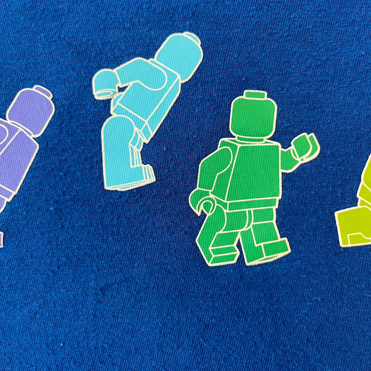 Lego Rainbow Minifigures Blue Tee from 2010, officially licensed by the Lego Group. Crafted from 100% cotton, it features seven colorful Lego minifigures in motion across the chest. Designed in a blue colorway with a small Lego tag on the left sleeve.