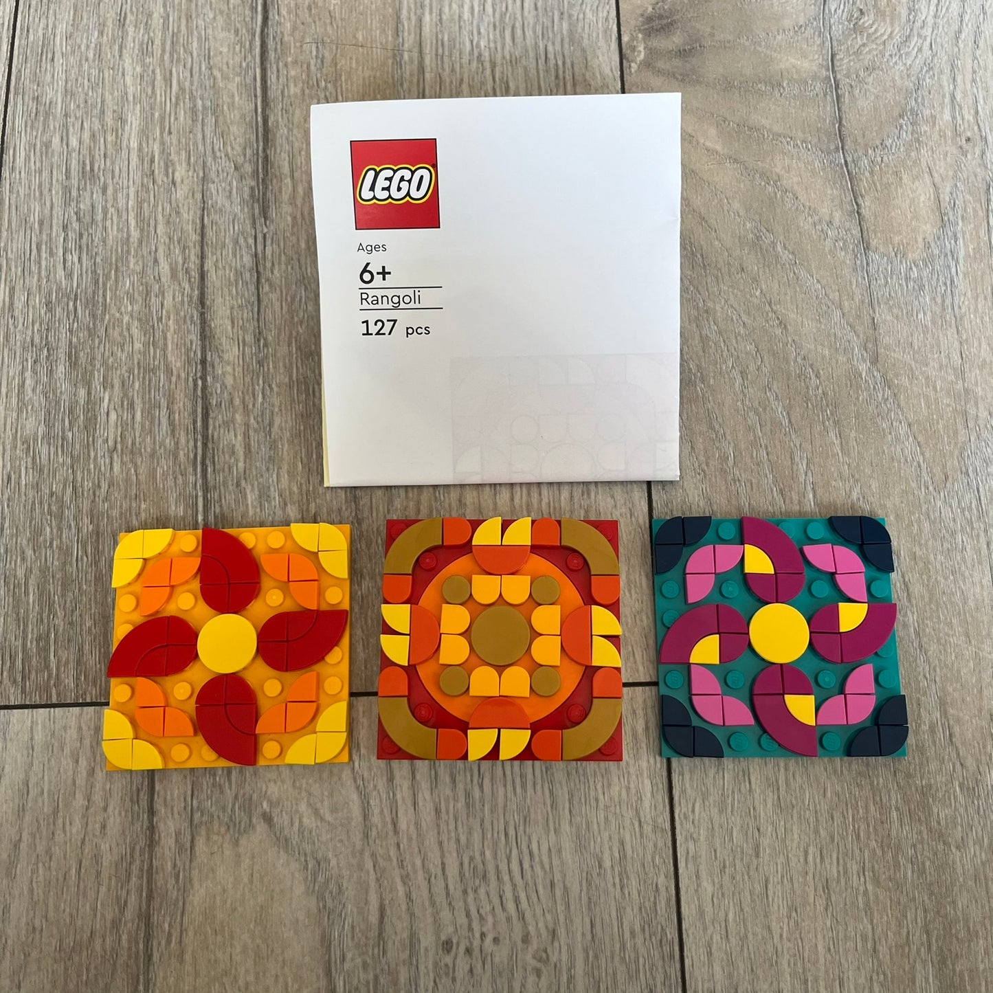 LEGO Rangoli Make and Take Build is a limited promotional LEGO set distributed as an exclusive “make & take” event and not available for retail purchase. The model features three small square rangoli-style builds arranged in decorative patterns. The designs use combinations of yellow, orange, red, brown, blue, and pink elements, with the first and third patterns matching each other.