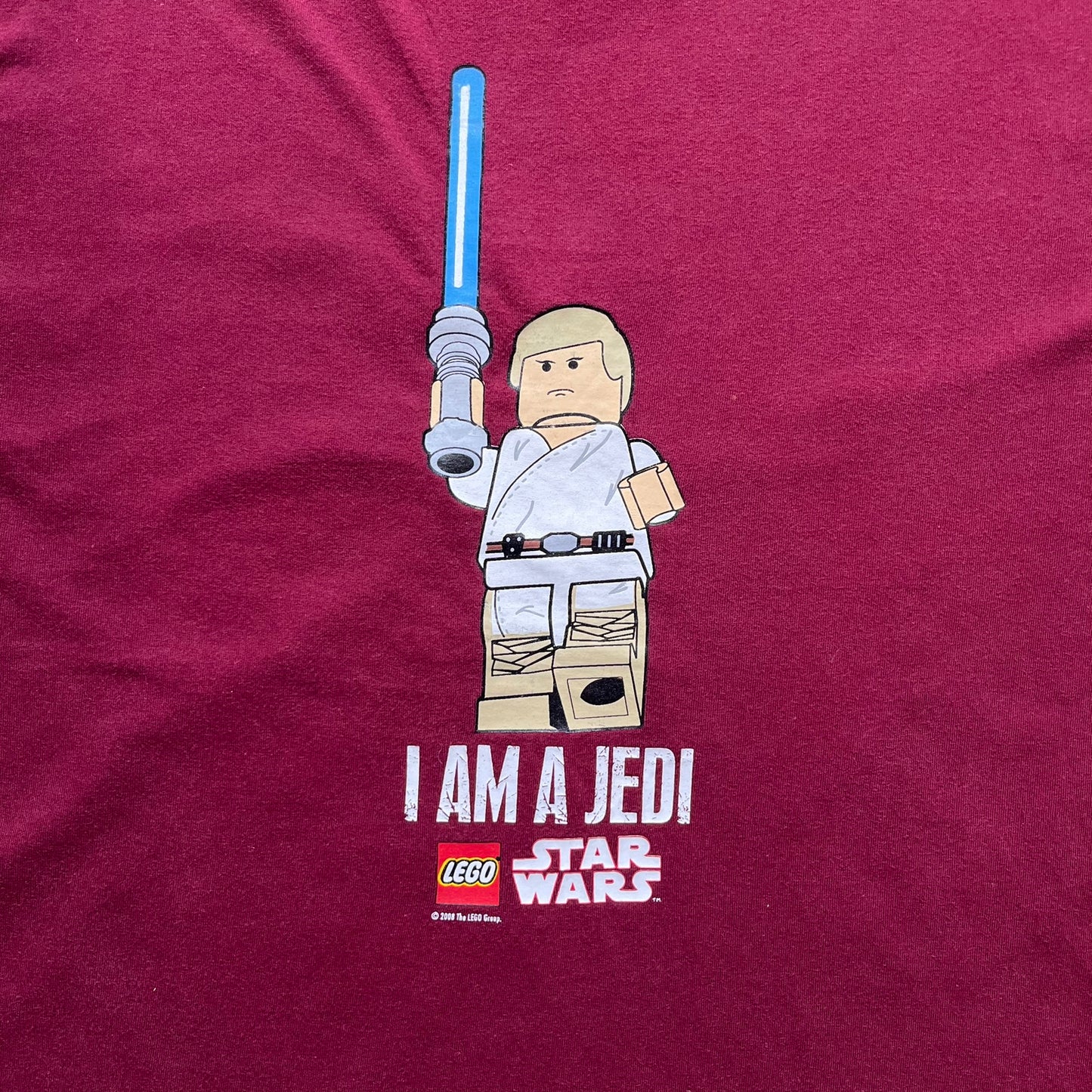 This Lego Star Wars 2008 Jedi Tee (Medium) in burgundy showcases Luke Skywalker wielding a blue lightsaber with a 'cracked' “I Am a Jedi” text. Officially licensed by Lego Star Wars, the tee includes authentic 2008 branding under the Luke minifigure graphic.