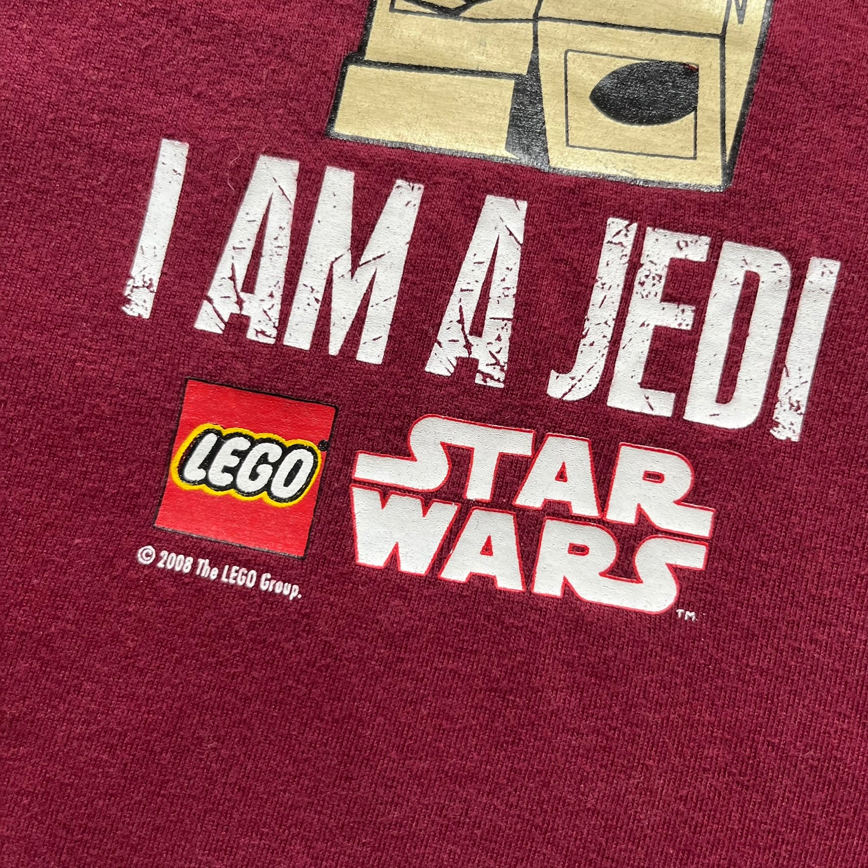 This Lego Star Wars 2008 Jedi Tee (Medium) in burgundy showcases Luke Skywalker wielding a blue lightsaber with a 'cracked' “I Am a Jedi” text. Officially licensed by Lego Star Wars, the tee includes authentic 2008 branding under the Luke minifigure graphic.