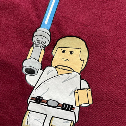 This Lego Star Wars 2008 Jedi Tee (Medium) in burgundy showcases Luke Skywalker wielding a blue lightsaber with a 'cracked' “I Am a Jedi” text. Officially licensed by Lego Star Wars, the tee includes authentic 2008 branding under the Luke minifigure graphic.