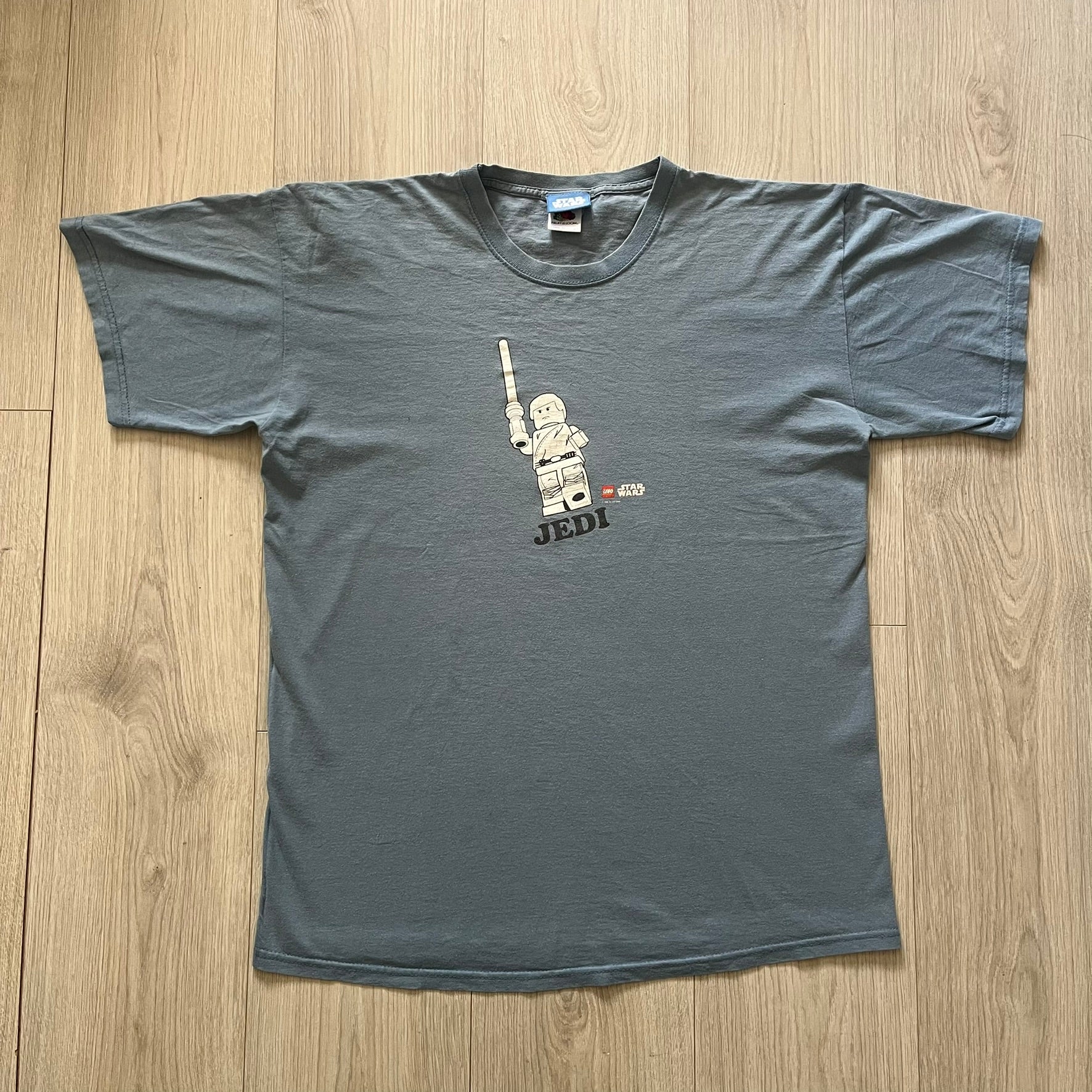 Vintage Lego Star Wars Jedi tee from 2008 in light blue. Showcases a clean white Lego Skywalker graphic with lightsaber and bold “Jedi” lettering underneath. Officially licensed and made on a soft Fruit of the Loom blank.