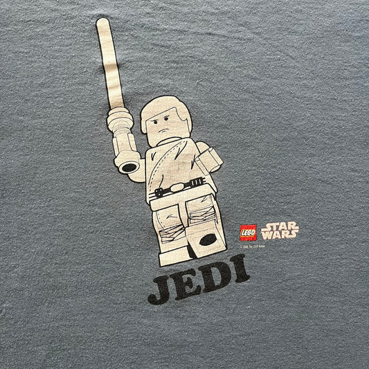 Vintage Lego Star Wars Jedi tee from 2008 in light blue. Showcases a clean white Lego Skywalker graphic with lightsaber and bold “Jedi” lettering underneath. Officially licensed and made on a soft Fruit of the Loom blank.