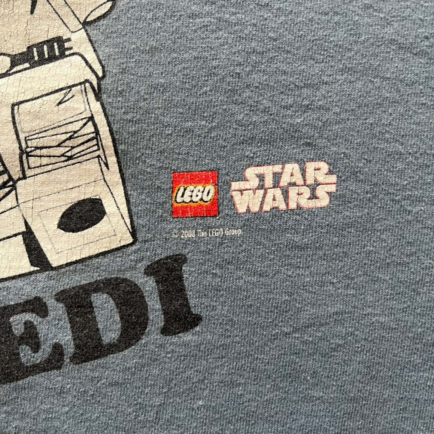 Vintage Lego Star Wars Jedi tee from 2008 in light blue. Showcases a clean white Lego Skywalker graphic with lightsaber and bold “Jedi” lettering underneath. Officially licensed and made on a soft Fruit of the Loom blank.