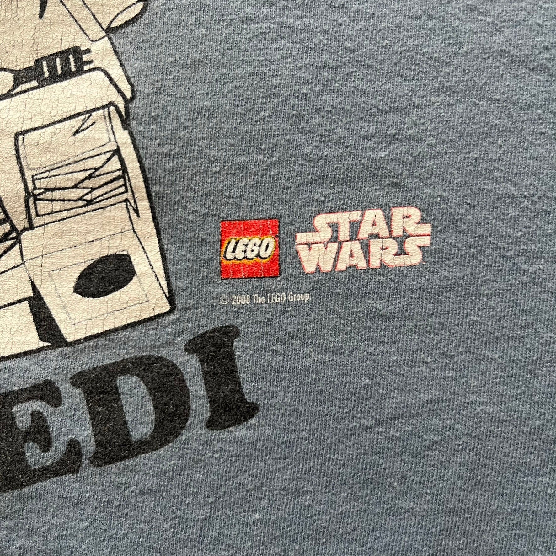 Vintage Lego Star Wars Jedi tee from 2008 in light blue. Showcases a clean white Lego Skywalker graphic with lightsaber and bold “Jedi” lettering underneath. Officially licensed and made on a soft Fruit of the Loom blank.