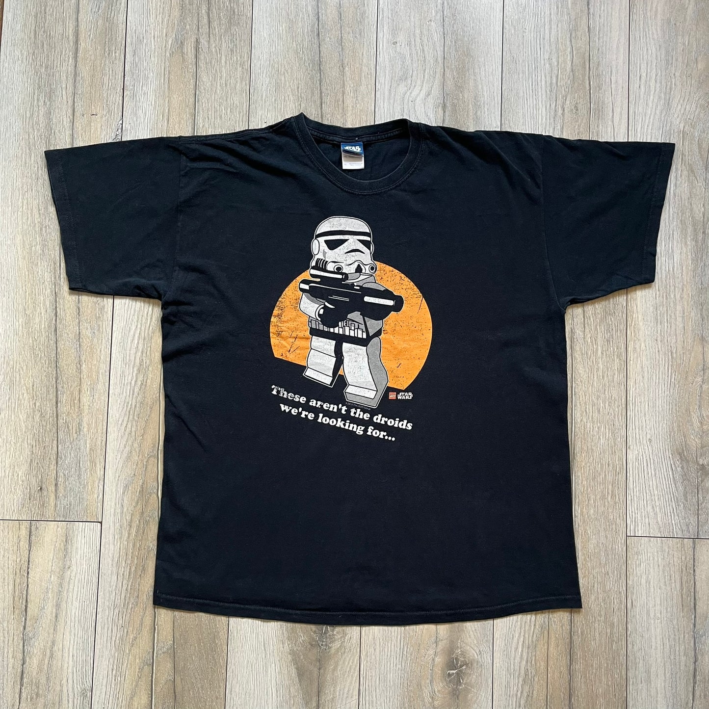 Lego Star Wars 2010 Stormtrooper Tee is a black T-shirt featuring a front graphic of a Lego Stormtrooper holding a blaster against an orange background. Below the graphic is the text “These aren’t the droids we’re looking for…” along with the Lego Star Wars logo and small 2010 licensing print.
