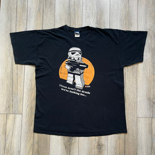 Lego Star Wars 2010 Stormtrooper Tee is a black T-shirt featuring a front graphic of a Lego Stormtrooper holding a blaster against an orange background. Below the graphic is the text “These aren’t the droids we’re looking for…” along with the Lego Star Wars logo and small 2010 licensing print.