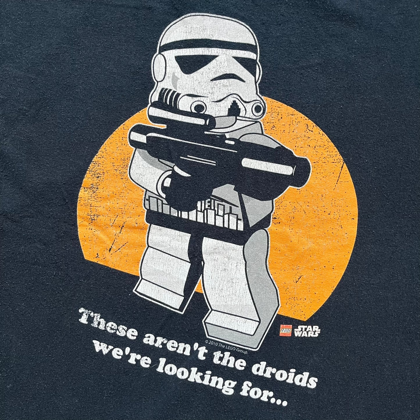 Lego Star Wars 2010 Stormtrooper Tee is a black T-shirt featuring a front graphic of a Lego Stormtrooper holding a blaster against an orange background. Below the graphic is the text “These aren’t the droids we’re looking for…” along with the Lego Star Wars logo and small 2010 licensing print.