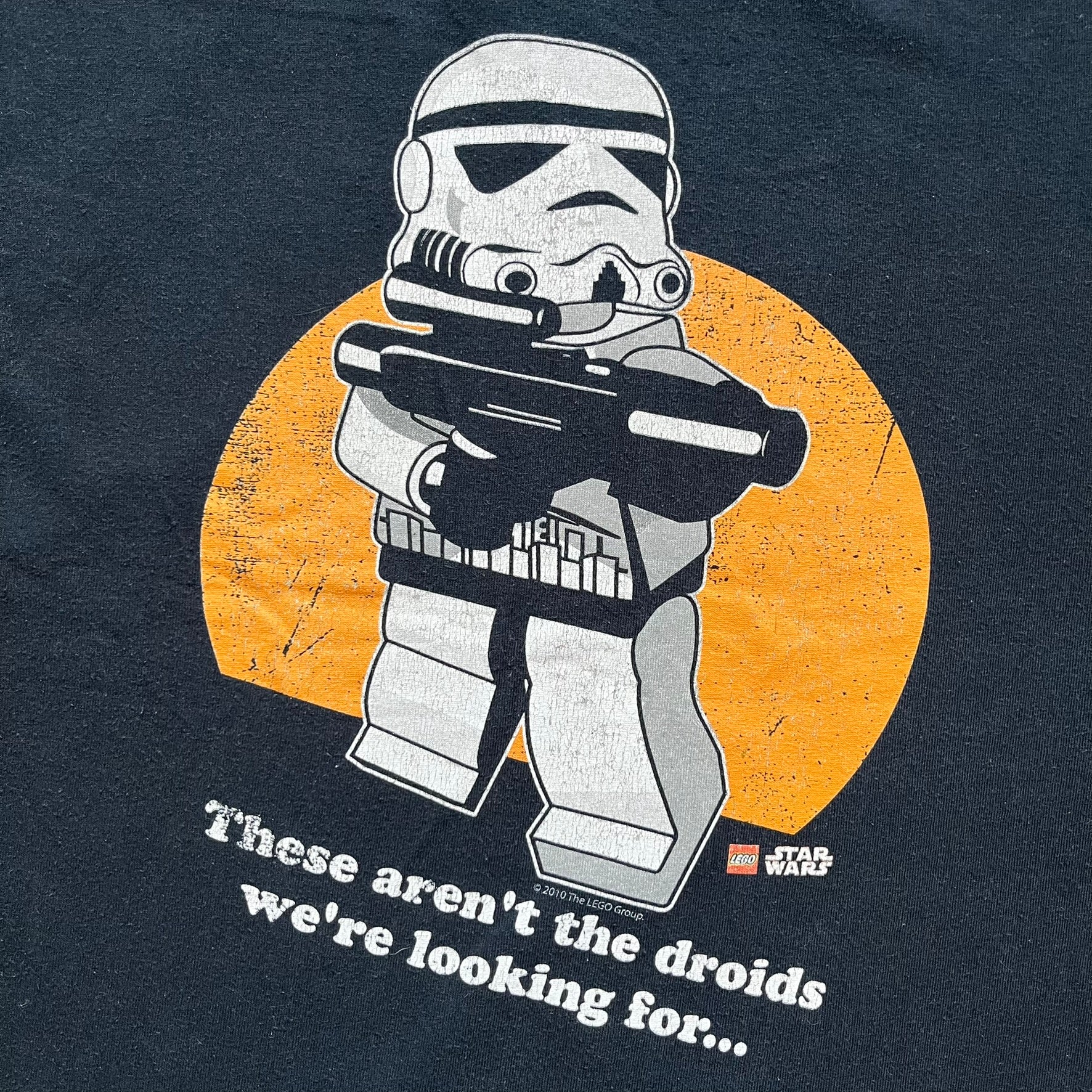 Lego Star Wars 2010 Stormtrooper Tee is a black T-shirt featuring a front graphic of a Lego Stormtrooper holding a blaster against an orange background. Below the graphic is the text “These aren’t the droids we’re looking for…” along with the Lego Star Wars logo and small 2010 licensing print.