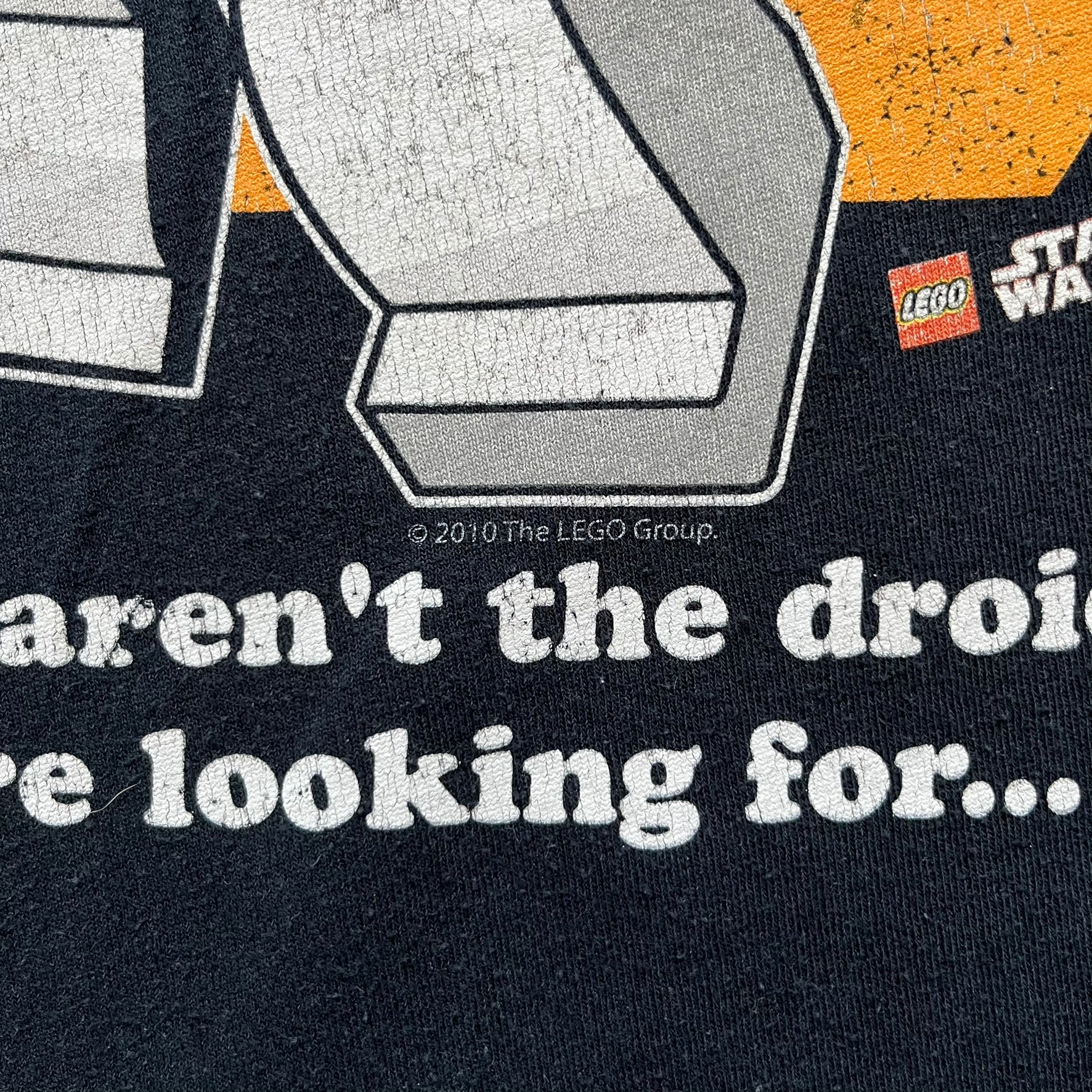 Lego Star Wars 2010 Stormtrooper Tee is a black T-shirt featuring a front graphic of a Lego Stormtrooper holding a blaster against an orange background. Below the graphic is the text “These aren’t the droids we’re looking for…” along with the Lego Star Wars logo and small 2010 licensing print.