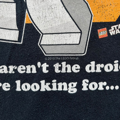 Lego Star Wars 2010 Stormtrooper Tee is a black T-shirt featuring a front graphic of a Lego Stormtrooper holding a blaster against an orange background. Below the graphic is the text “These aren’t the droids we’re looking for…” along with the Lego Star Wars logo and small 2010 licensing print.