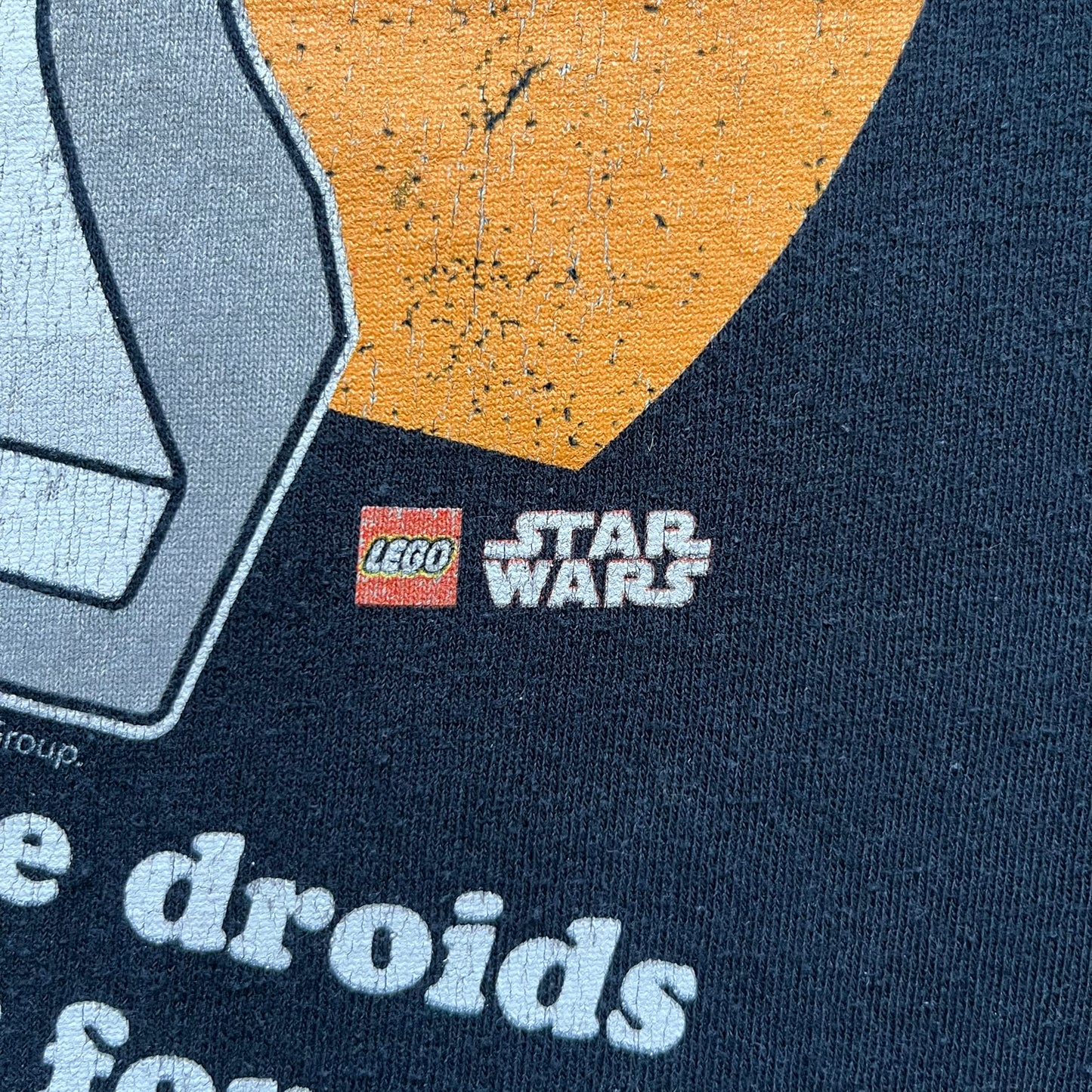 Lego Star Wars 2010 Stormtrooper Tee is a black T-shirt featuring a front graphic of a Lego Stormtrooper holding a blaster against an orange background. Below the graphic is the text “These aren’t the droids we’re looking for…” along with the Lego Star Wars logo and small 2010 licensing print.