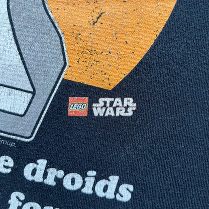 Lego Star Wars 2010 Stormtrooper Tee is a black T-shirt featuring a front graphic of a Lego Stormtrooper holding a blaster against an orange background. Below the graphic is the text “These aren’t the droids we’re looking for…” along with the Lego Star Wars logo and small 2010 licensing print.