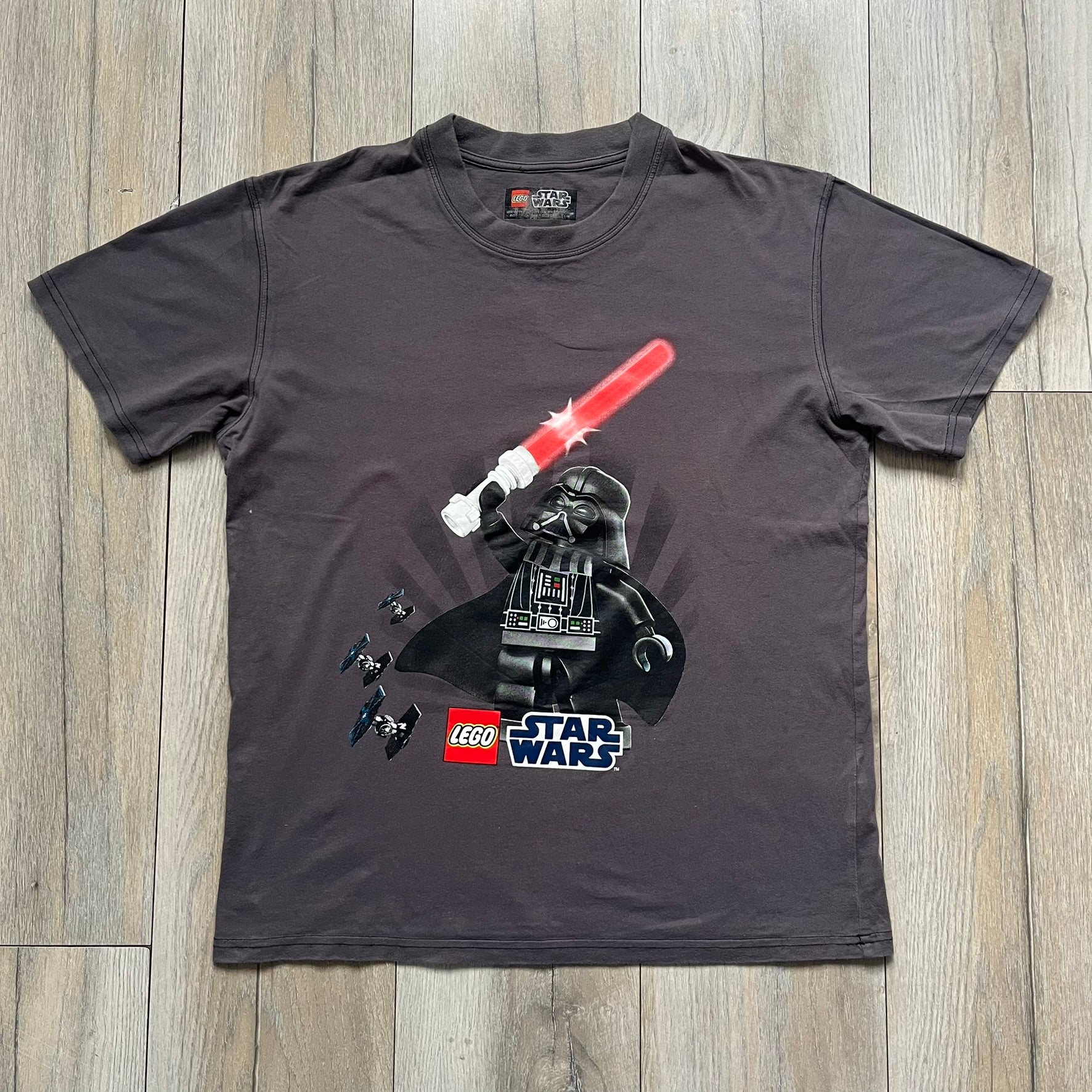 LEGO Star Wars 2012 Darth Vader Tee is a gray-brown cotton T-shirt featuring a large front graphic of Darth Vader holding a red lightsaber in a triumphant pose. The design includes three small TIE fighters flying beside him, with the LEGO Star Wars logo printed underneath. The shirt includes a 2012 licensed neck tag and official Star Wars branding.