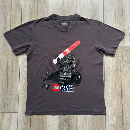 LEGO Star Wars 2012 Darth Vader Tee is a gray-brown cotton T-shirt featuring a large front graphic of Darth Vader holding a red lightsaber in a triumphant pose. The design includes three small TIE fighters flying beside him, with the LEGO Star Wars logo printed underneath. The shirt includes a 2012 licensed neck tag and official Star Wars branding.