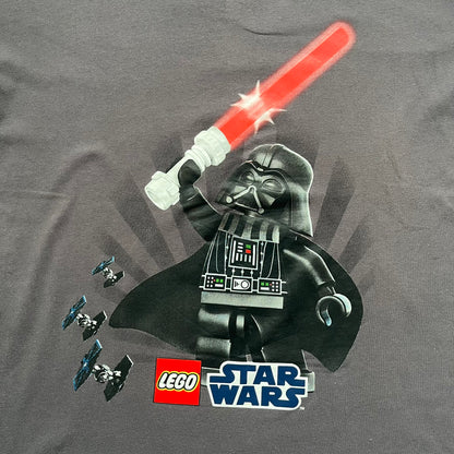 LEGO Star Wars 2012 Darth Vader Tee is a gray-brown cotton T-shirt featuring a large front graphic of Darth Vader holding a red lightsaber in a triumphant pose. The design includes three small TIE fighters flying beside him, with the LEGO Star Wars logo printed underneath. The shirt includes a 2012 licensed neck tag and official Star Wars branding.