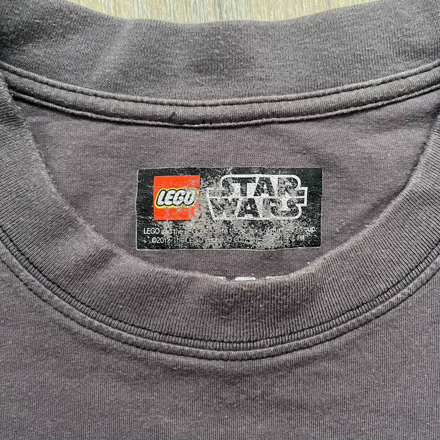 LEGO Star Wars 2012 Darth Vader Tee is a gray-brown cotton T-shirt featuring a large front graphic of Darth Vader holding a red lightsaber in a triumphant pose. The design includes three small TIE fighters flying beside him, with the LEGO Star Wars logo printed underneath. The shirt includes a 2012 licensed neck tag and official Star Wars branding.