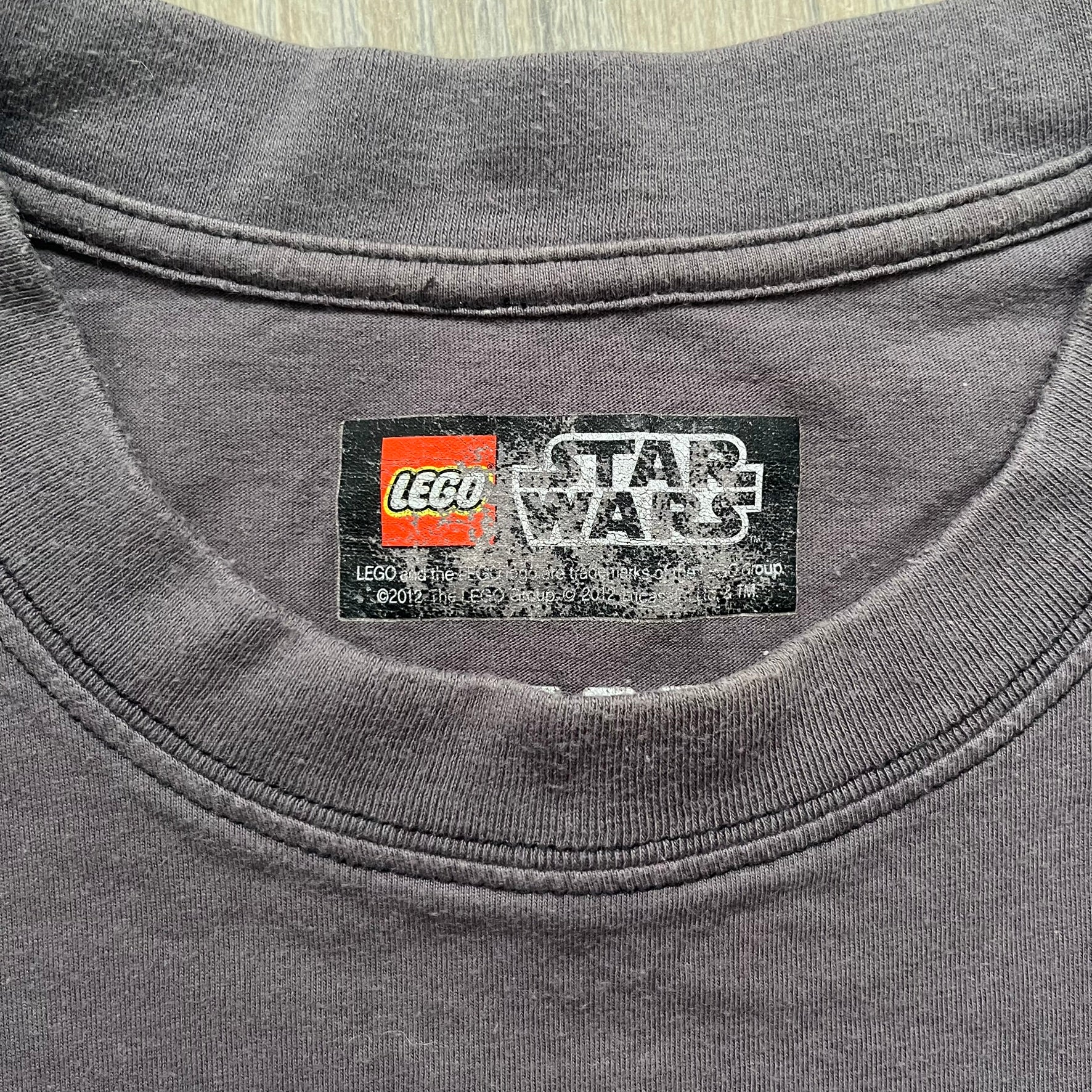 LEGO Star Wars 2012 Darth Vader Tee is a gray-brown cotton T-shirt featuring a large front graphic of Darth Vader holding a red lightsaber in a triumphant pose. The design includes three small TIE fighters flying beside him, with the LEGO Star Wars logo printed underneath. The shirt includes a 2012 licensed neck tag and official Star Wars branding.