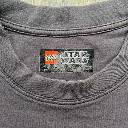 LEGO Star Wars 2012 Darth Vader Tee is a gray-brown cotton T-shirt featuring a large front graphic of Darth Vader holding a red lightsaber in a triumphant pose. The design includes three small TIE fighters flying beside him, with the LEGO Star Wars logo printed underneath. The shirt includes a 2012 licensed neck tag and official Star Wars branding.
