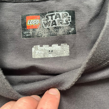 LEGO Star Wars 2012 Darth Vader Tee is a gray-brown cotton T-shirt featuring a large front graphic of Darth Vader holding a red lightsaber in a triumphant pose. The design includes three small TIE fighters flying beside him, with the LEGO Star Wars logo printed underneath. The shirt includes a 2012 licensed neck tag and official Star Wars branding.