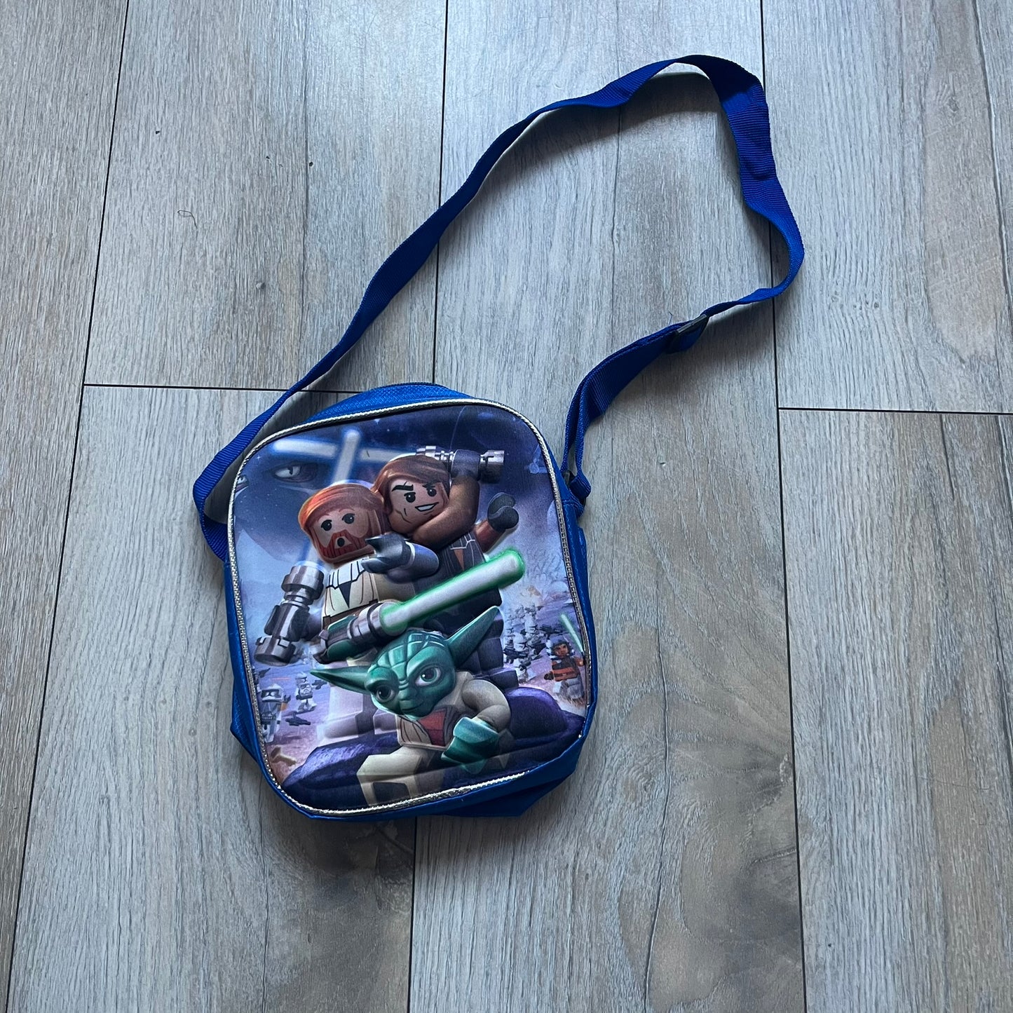 Lego Star Wars III: The Clone Wars Shoulder Bag is a blue shoulder bag featuring a raised 3D front graphic panel (not printed) with Lego Star Wars artwork. The design shows Obi-Wan Kenobi, Anakin Skywalker, and Yoda with lightsabers on a battlefield scene, with clone troopers, Ahsoka Tano, and General Grievous in the background.