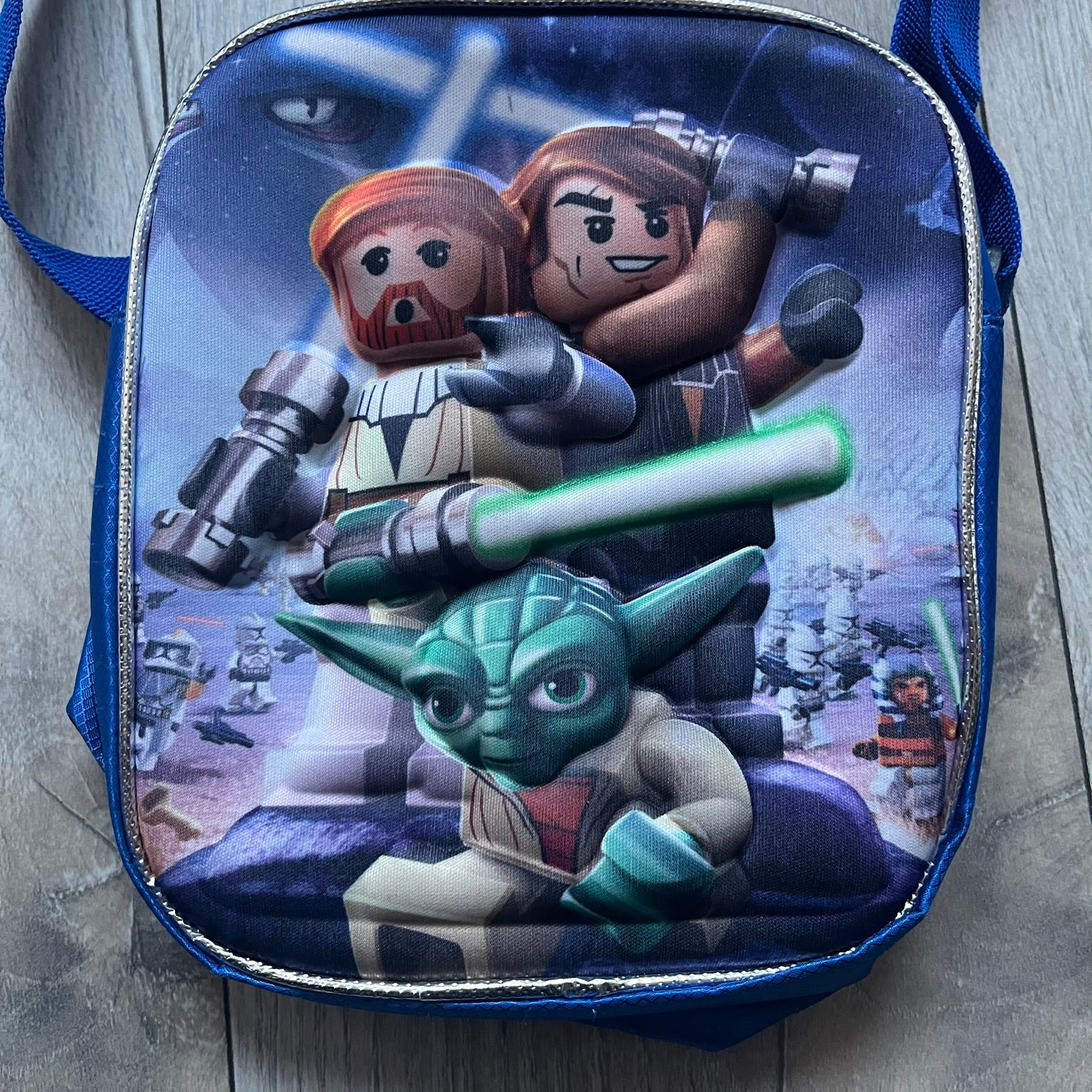 Lego Star Wars III: The Clone Wars Shoulder Bag is a blue shoulder bag featuring a raised 3D front graphic panel (not printed) with Lego Star Wars artwork. The design shows Obi-Wan Kenobi, Anakin Skywalker, and Yoda with lightsabers on a battlefield scene, with clone troopers, Ahsoka Tano, and General Grievous in the background.