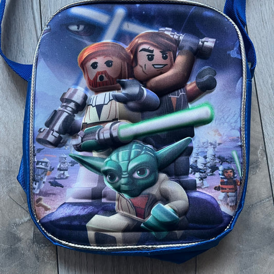 Lego Star Wars III: The Clone Wars Shoulder Bag is a blue shoulder bag featuring a raised 3D front graphic panel (not printed) with Lego Star Wars artwork. The design shows Obi-Wan Kenobi, Anakin Skywalker, and Yoda with lightsabers on a battlefield scene, with clone troopers, Ahsoka Tano, and General Grievous in the background.