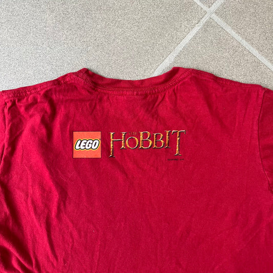 Lego The Hobbit 2014 Press Kit Red Tee features a red T-shirt with a full-front graphic designed to resemble Bilbo Baggins’ outfit in LEGO form, including a green undershirt, beige tie, and dark details. The upper back has a LEGO The Hobbit logo with 2014 licensing text. Originally included as part of a press kit with additional items.