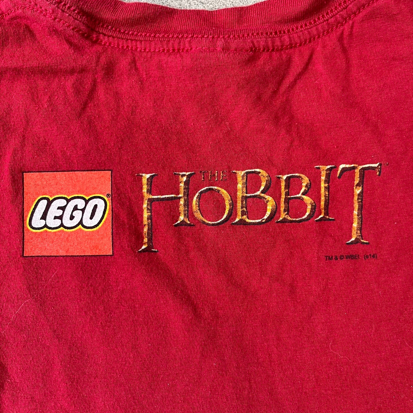 Lego The Hobbit 2014 Press Kit Red Tee features a red T-shirt with a full-front graphic designed to resemble Bilbo Baggins’ outfit in LEGO form, including a green undershirt, beige tie, and dark details. The upper back has a LEGO The Hobbit logo with 2014 licensing text. Originally included as part of a press kit with additional items.