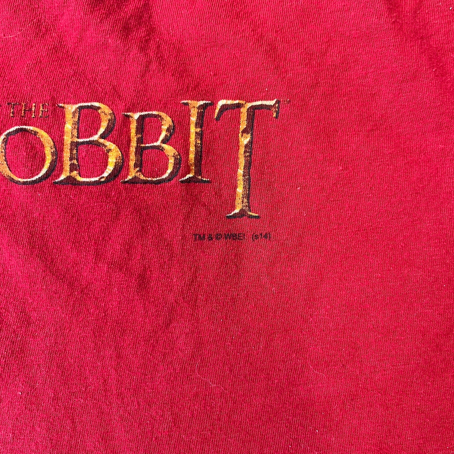 Lego The Hobbit 2014 Press Kit Red Tee features a red T-shirt with a full-front graphic designed to resemble Bilbo Baggins’ outfit in LEGO form, including a green undershirt, beige tie, and dark details. The upper back has a LEGO The Hobbit logo with 2014 licensing text. Originally included as part of a press kit with additional items.