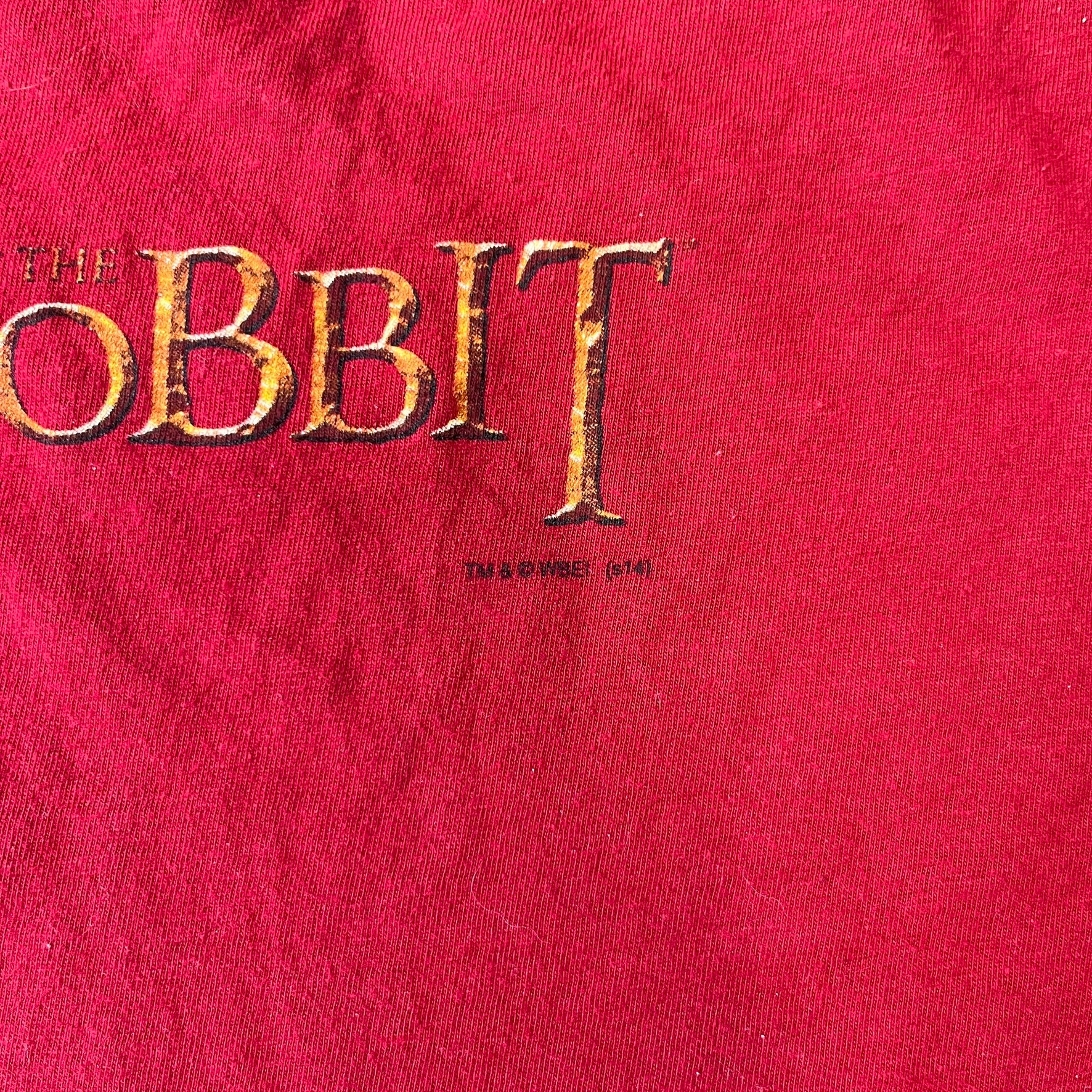 Lego The Hobbit 2014 Press Kit Red Tee features a red T-shirt with a full-front graphic designed to resemble Bilbo Baggins’ outfit in LEGO form, including a green undershirt, beige tie, and dark details. The upper back has a LEGO The Hobbit logo with 2014 licensing text. Originally included as part of a press kit with additional items.