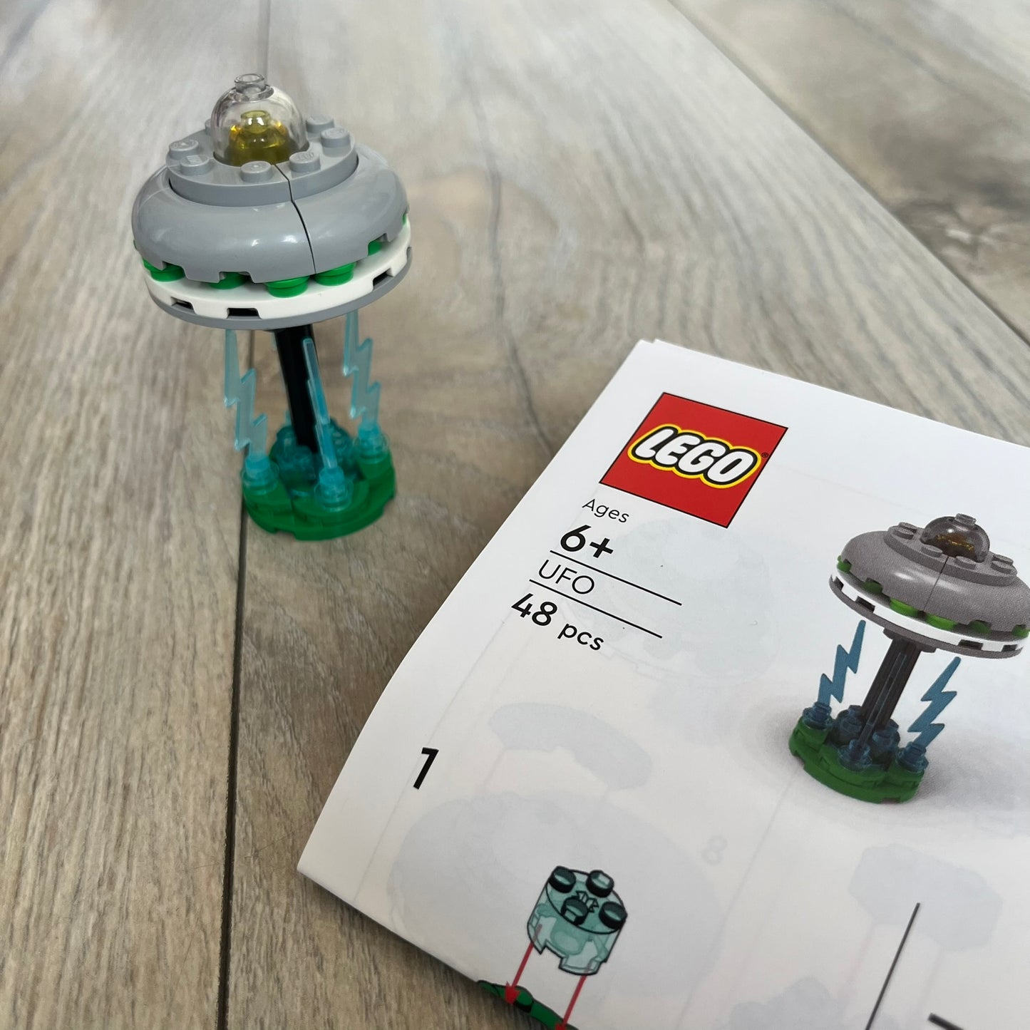 LEGO UFO Make and Take Build is a limited promotional LEGO set distributed as an exclusive “make & take” event and not available for retail purchase. The model features a small grey UFO with a black underside, displayed on a green grass base. Blue lightning bolt elements extend downward from the UFO, representing beam-like lights.