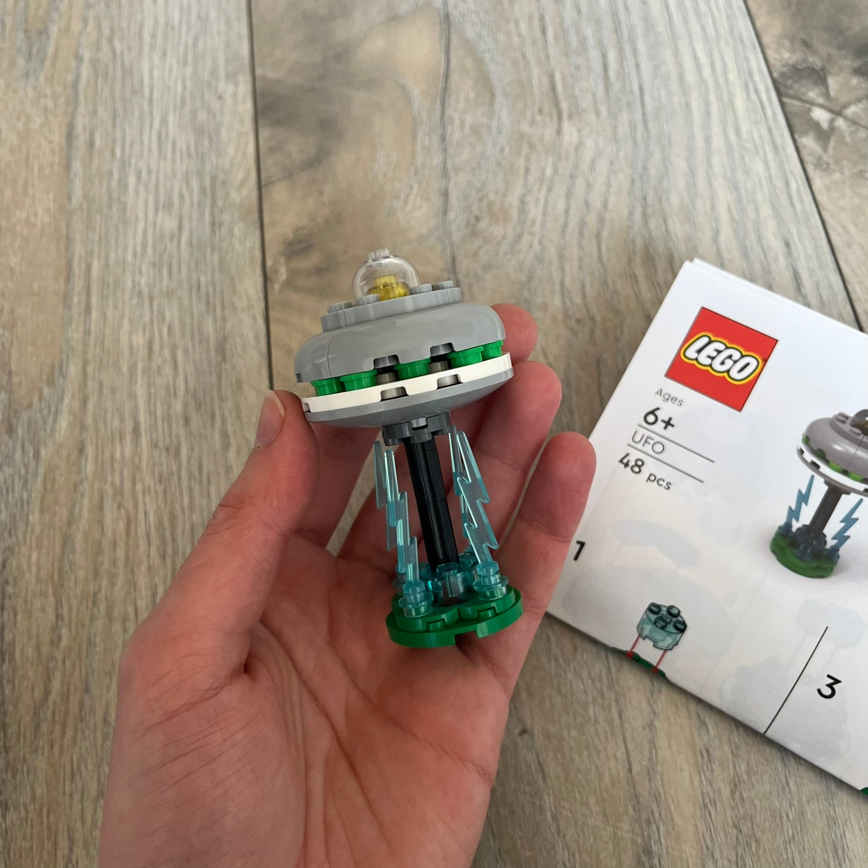 LEGO UFO Make and Take Build is a limited promotional LEGO set distributed as an exclusive “make & take” event and not available for retail purchase. The model features a small grey UFO with a black underside, displayed on a green grass base. Blue lightning bolt elements extend downward from the UFO, representing beam-like lights.