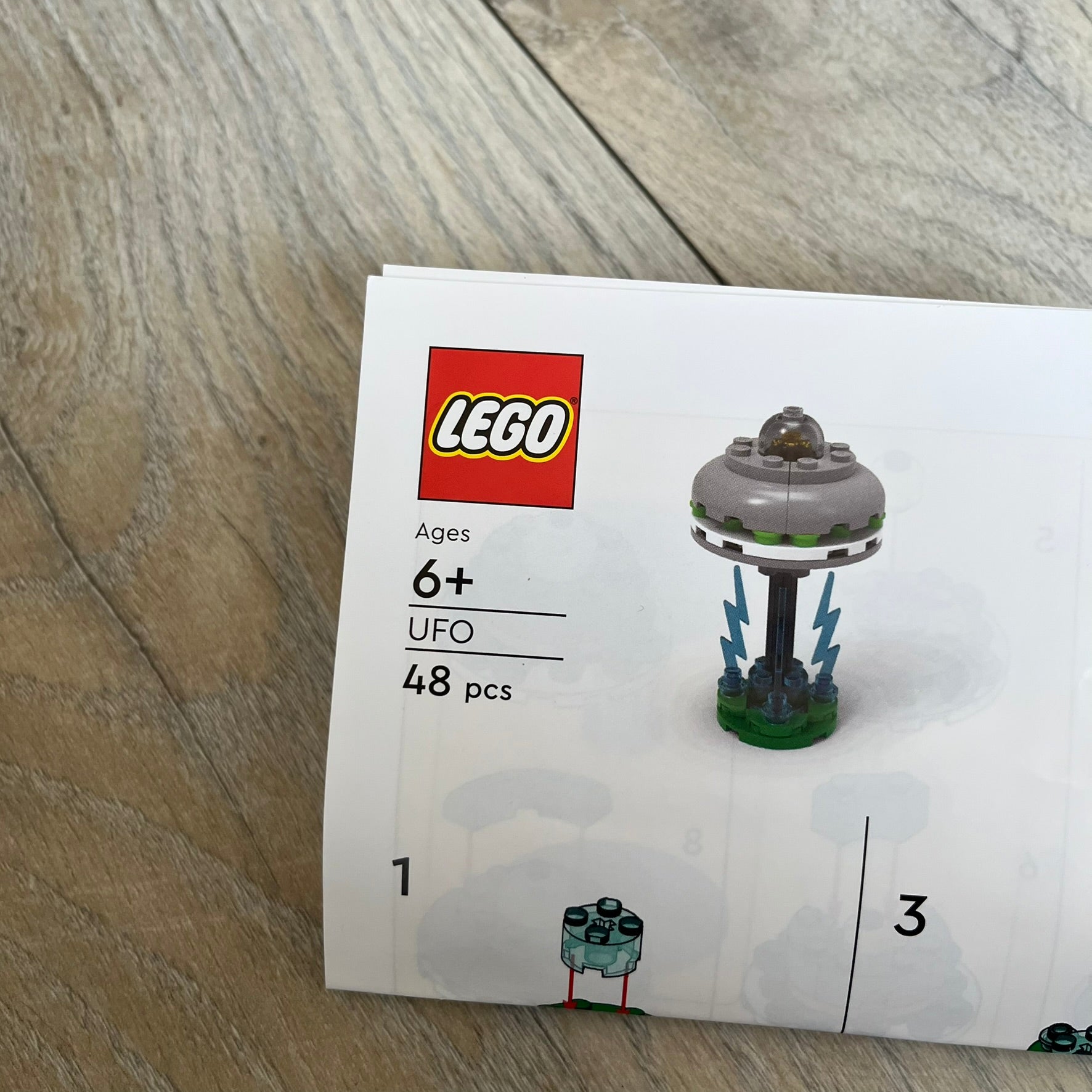 LEGO UFO Make and Take Build is a limited promotional LEGO set distributed as an exclusive “make & take” event and not available for retail purchase. The model features a small grey UFO with a black underside, displayed on a green grass base. Blue lightning bolt elements extend downward from the UFO, representing beam-like lights.