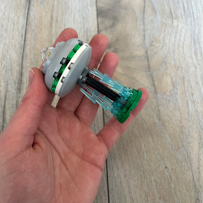 LEGO UFO Make and Take Build is a limited promotional LEGO set distributed as an exclusive “make & take” event and not available for retail purchase. The model features a small grey UFO with a black underside, displayed on a green grass base. Blue lightning bolt elements extend downward from the UFO, representing beam-like lights.