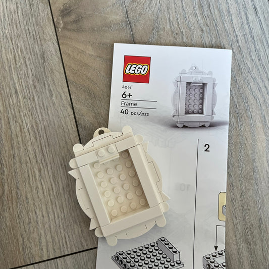 LEGO White Frame Make and Take Build is a limited promotional LEGO set distributed as an exclusive “make & take” event and not available for retail purchase. The model features a fully white 3D frame styled like a picture frame, designed for use as a photo holder. The back includes a hole for hanging or display.