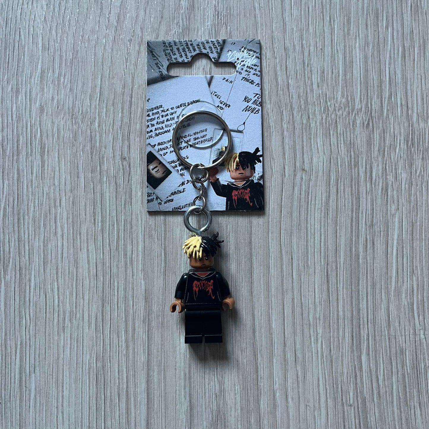 Lego XXXTentacion “XXXBlocktentacion” keychain featuring a custom Lego figure of XXXTentacion in a black/red revenge hoodie, with his classic split black and beige dreads in 3D. The figure is attached to a Lego-style recreation of the 2017 '17' album cover, reimagined in brick graphics. A great tribute and a creative collectible that merges music culture with fan-made Lego artistry, made by TheCanvasDon.