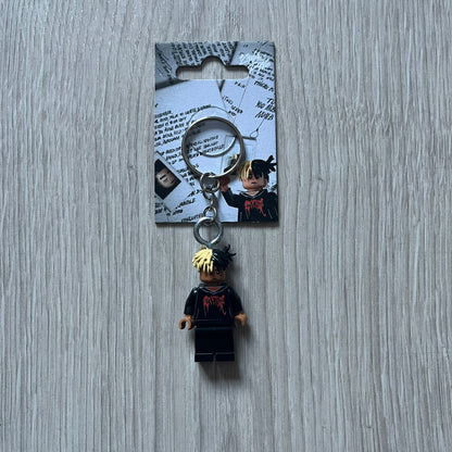 Lego XXXTentacion “XXXBlocktentacion” keychain featuring a custom Lego figure of XXXTentacion in a black/red revenge hoodie, with his classic split black and beige dreads in 3D. The figure is attached to a Lego-style recreation of the 2017 '17' album cover, reimagined in brick graphics. A great tribute and a creative collectible that merges music culture with fan-made Lego artistry, made by TheCanvasDon.