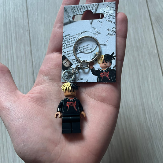 Lego XXXTentacion “XXXBlocktentacion” keychain featuring a custom Lego figure of XXXTentacion in a black/red revenge hoodie, with his classic split black and beige dreads in 3D. The figure is attached to a Lego-style recreation of the 2017 '17' album cover, reimagined in brick graphics. A great tribute and a creative collectible that merges music culture with fan-made Lego artistry, made by TheCanvasDon.